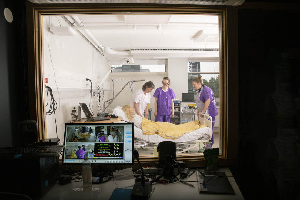 Tre personer står över en docka i en sjukhussäng, sett från ett annat rum i en simuleingsmiljö. Foto