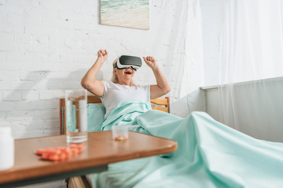 Person sitter på sjukhussäng med armarna i luften med VR-glasögon på sig. Foto. 