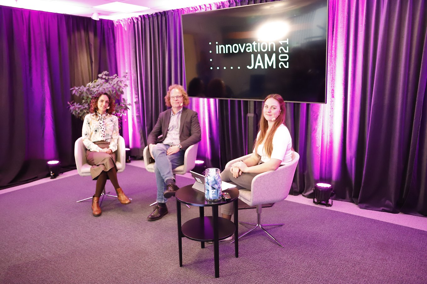 tre personer sitter i fåtöljer på en scen. Det är lila ljus bakom dem och en tv-skärm med texten Innovation Jam 2021. 